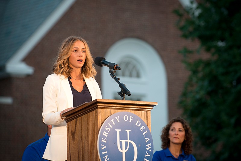 Nikki Dombrowski speaks at the 2016 Twilight Induction Ceremony. She is standing at a podium with the University of Delaware logo on it.