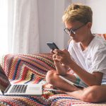 Preteen boy staring at cell phone with laptop open next to him.