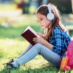 A girl reads a book under a tree with headphones on.
