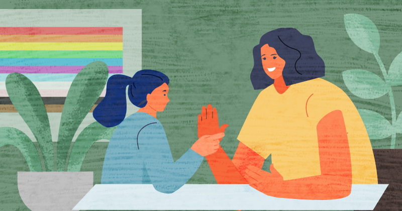 An adolescent student works with a teacher individually at a table with a rainbow flag behind them.