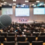 Image of a microphone in an auditorium with an audience and panel discussion.