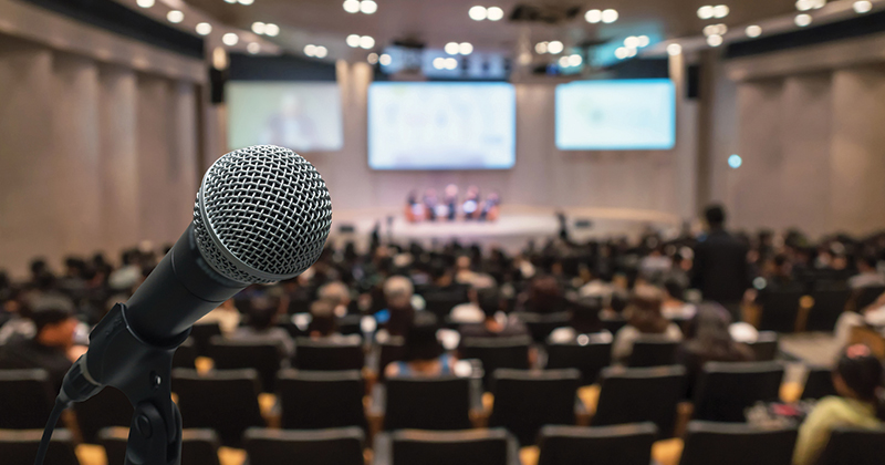 Image of a microphone in an auditorium with an audience and panel discussion.