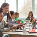 Children work with a teacher using computers in a classroom setting.