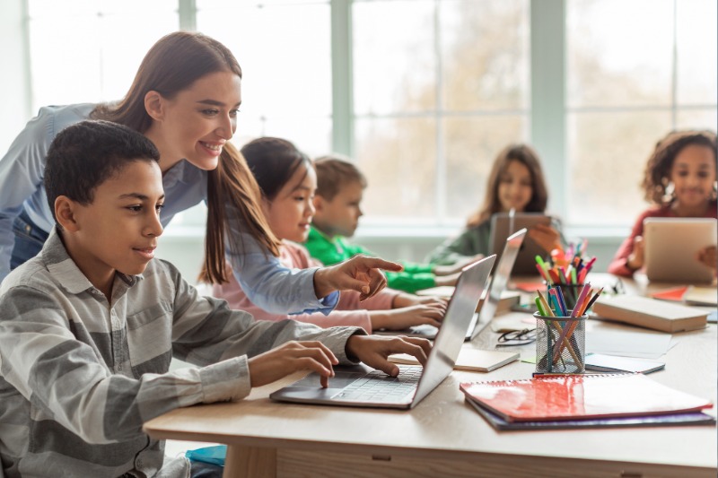 Children work with a teacher using computers in a classroom setting.