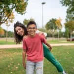 An adolescent boy playfully carries an older sister on his back in a public park.