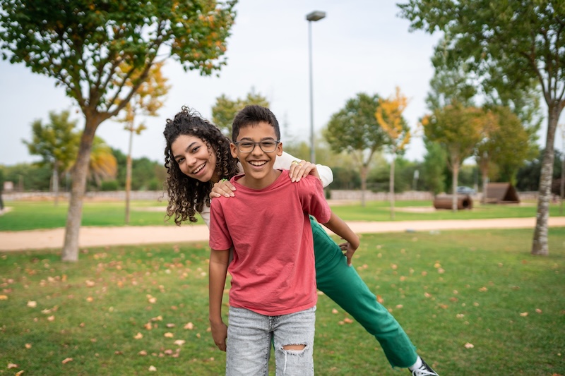 An adolescent boy playfully carries an older sister on his back in a public park.